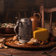 A wedge of cheese and pewter tankard sit on a tray on a crumb-covered wooden table.